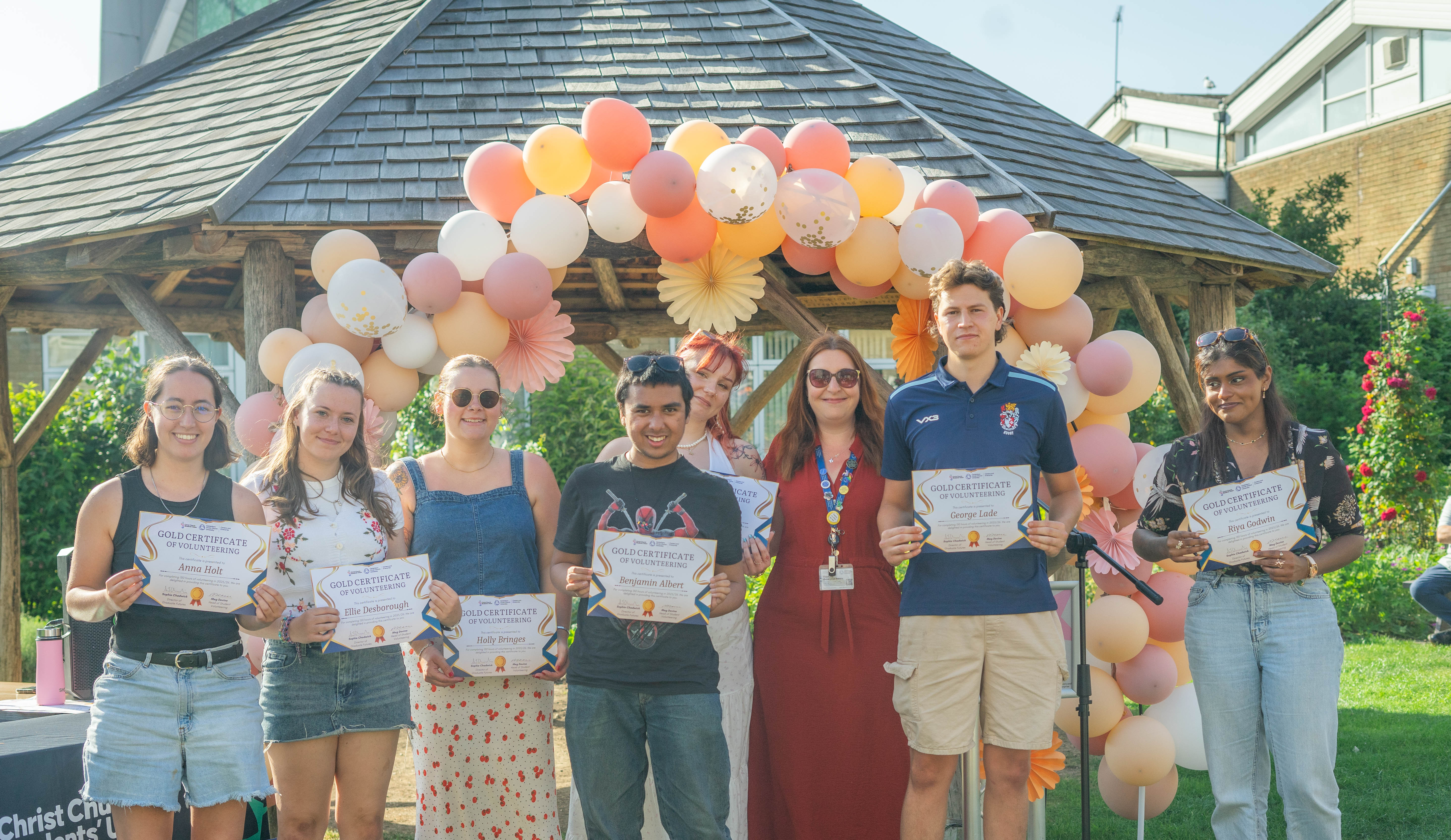 a collection of volunteers standing outside under a balloon arch holding their certificates