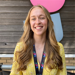 White person with long wavey mousey brown hair, wearing a yellow top and smiling at the camera.