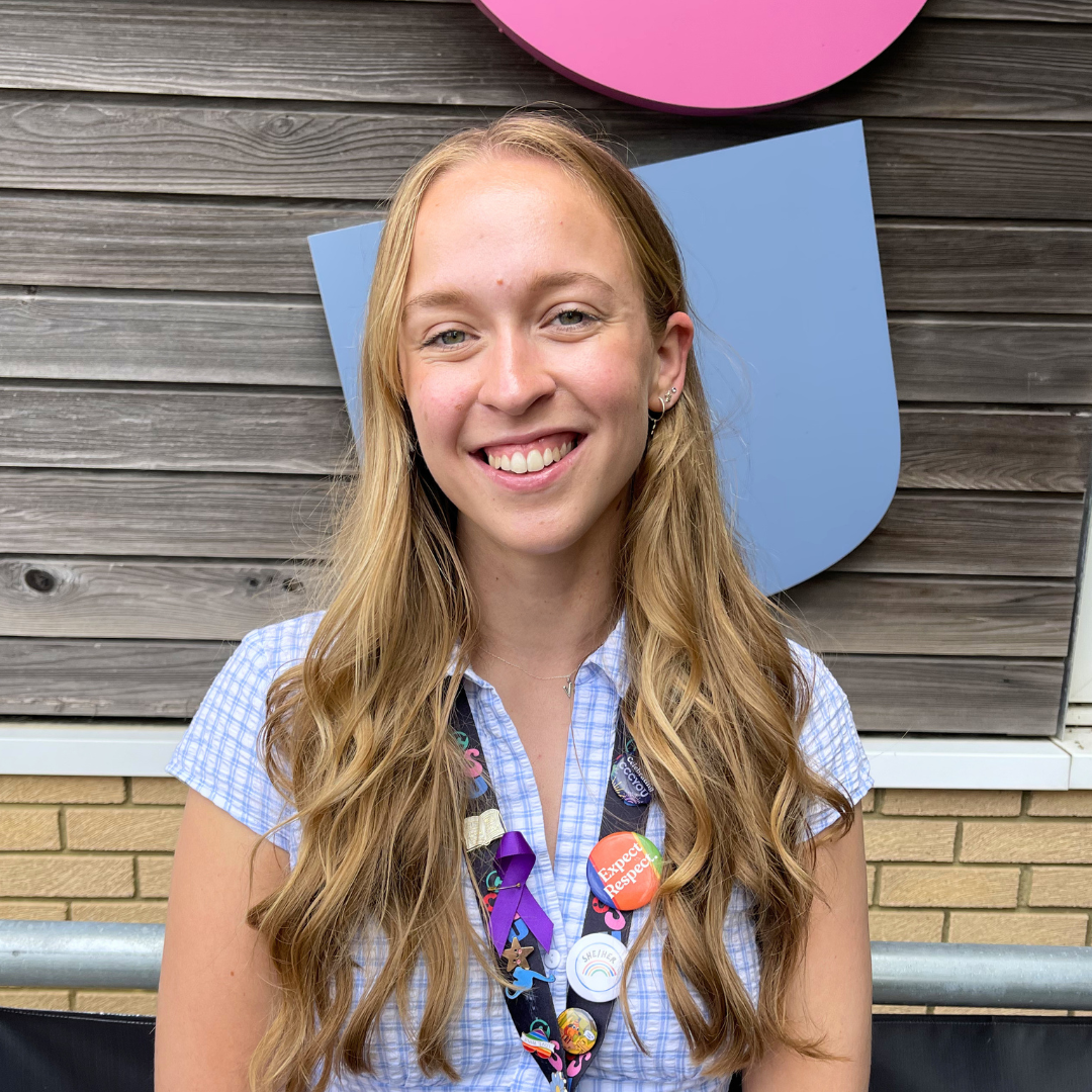White person with long wavey mousey brown hair, wearing a blue top and smiling at the camera.