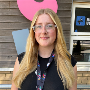 White person with long blonde hair, clear glasses wearing a black top smiling at the camera.