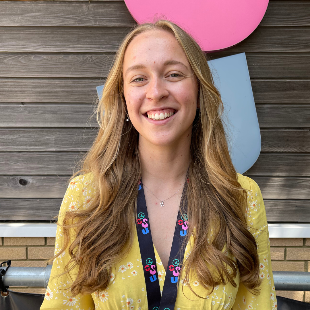 Portrait of person with long light brown hair, wearing a yellow top smiling at the camera