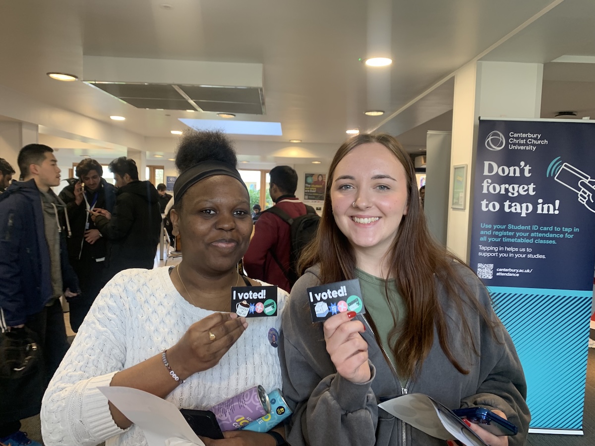Two female students holding up cards that read 'I voted' smiling at the camera