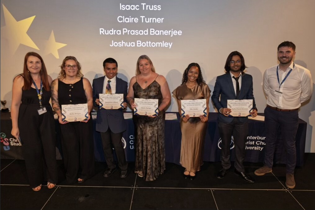 Group of 7 people stood on a stage at an award ceremony holding a certificate